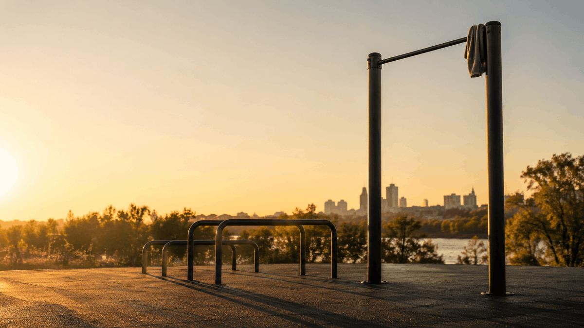 Empty outdoor pull-up bar and low parallel bars on rubberized ground at golden hour, with a folded towel over one bar end and long shadows