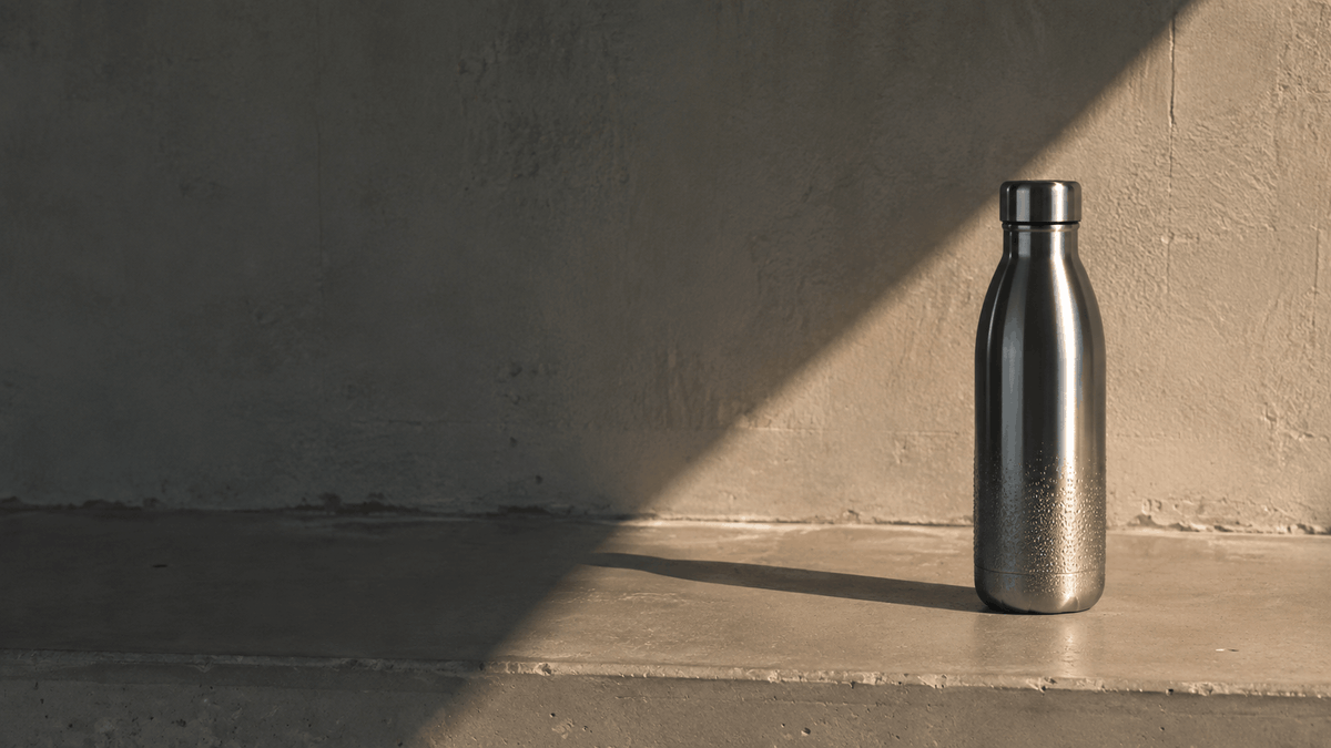 Unlabeled stainless steel water bottle standing on a bare concrete ledge, lit by a single band of low sunlight casting one long shadow