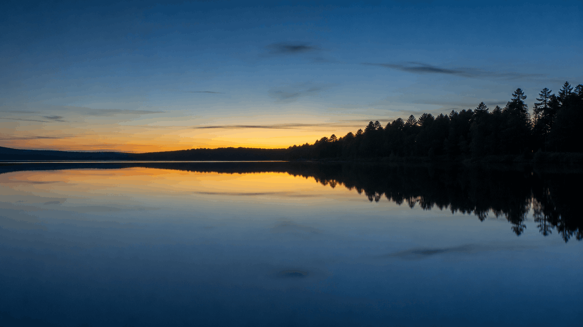 Still, mirror-flat lake surface at dusk, with a low tree line and the last warm band of sky reflected across the water and no ripples