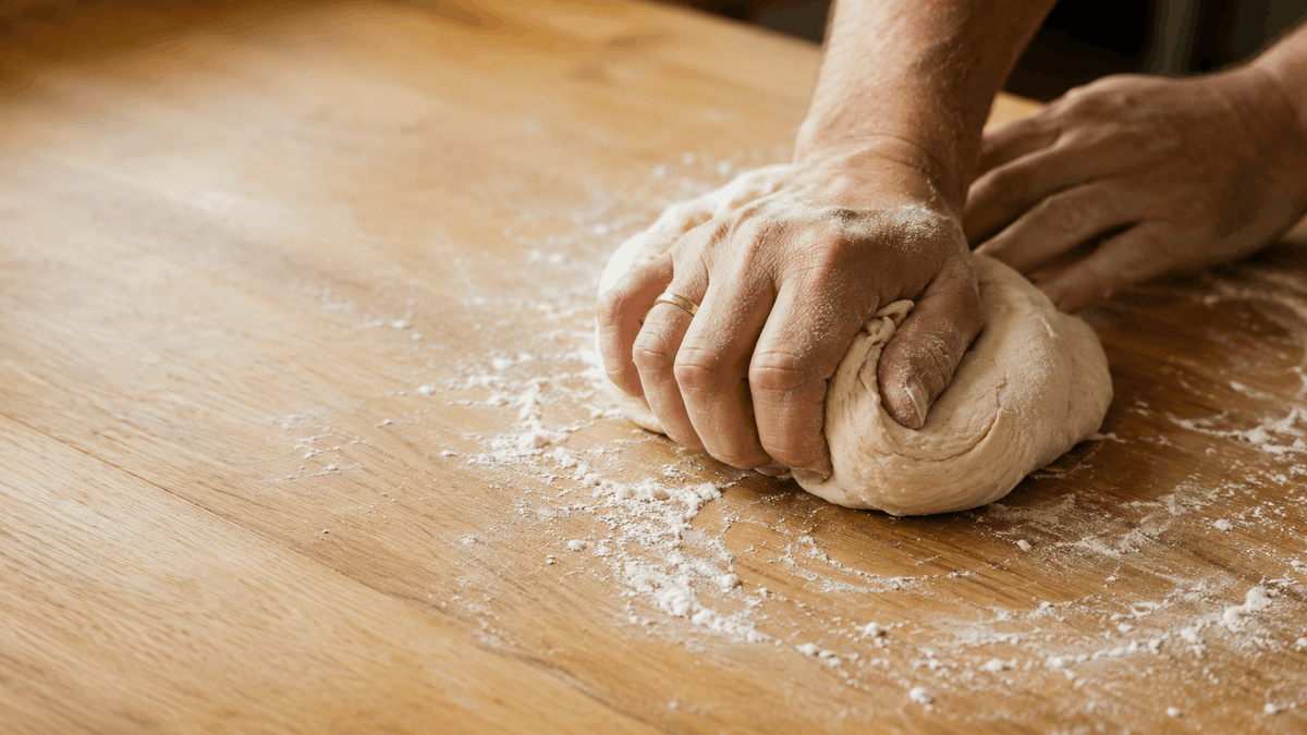 Hands kneading bread dough on a floured wooden board in warm daylight