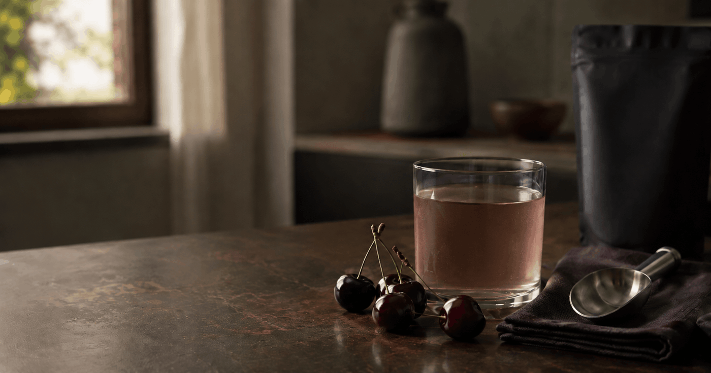 A clear glass of prepared black cherry flavored drink with dark cherries, a stainless scoop, and a neutral container on a dark wood surface.