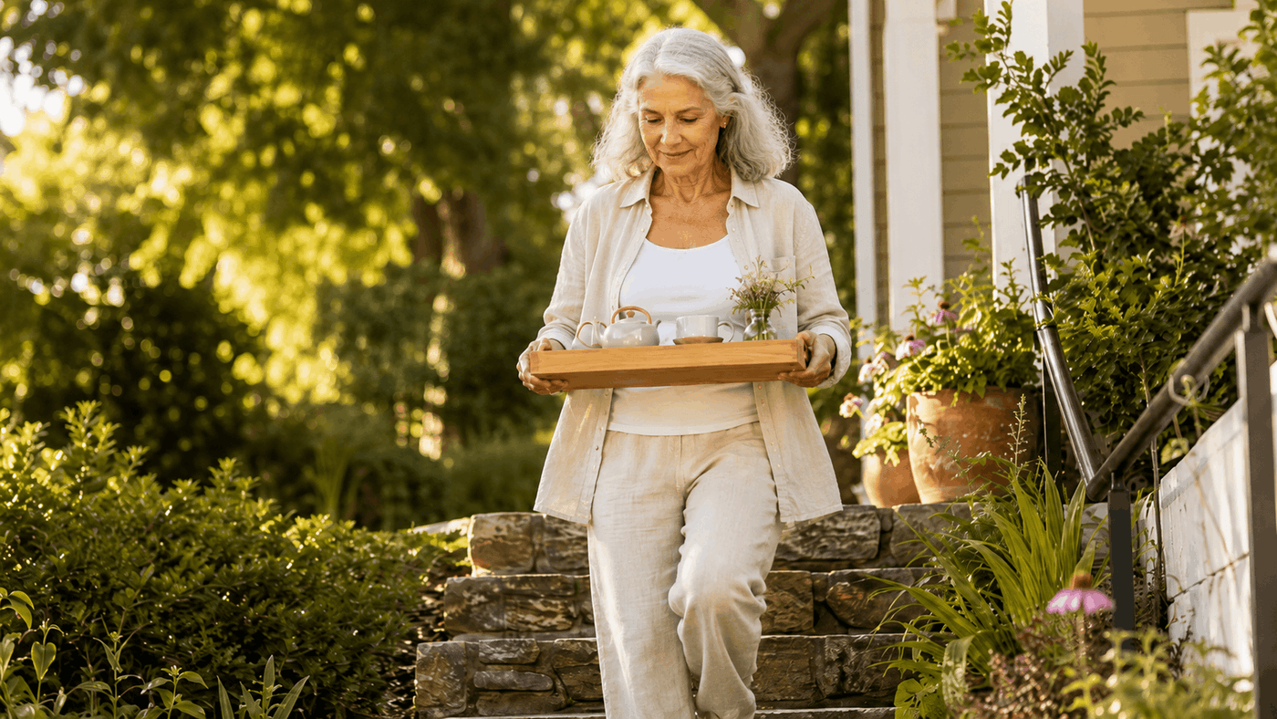 An active adult in her 60s confidently descending a few outdoor stesp with hands full (a tea tray), sure-footed, in a warm natural light.
