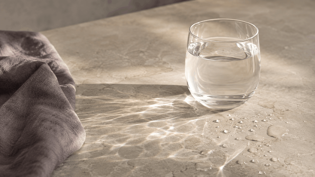A clear glass of water on a pale stone surface in warm morning light, with water droplets and soft violet-grey linen nearby.